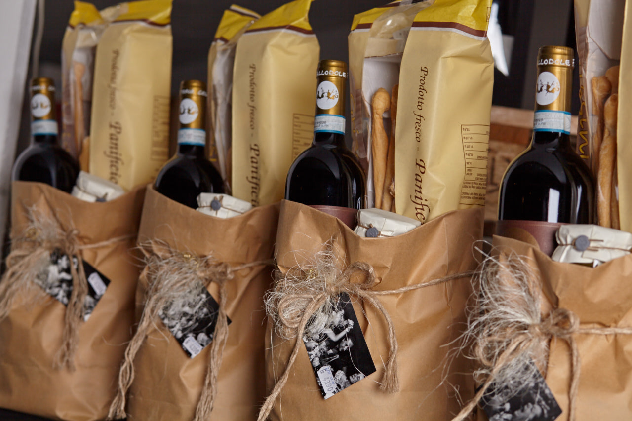 Geschenkpakete mit italienischer Feinkost – Weinflaschen, Grissini und Delikatessen in braunen Papiertüten mit Kordel und Anhänger
